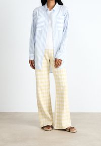 Světle modrá pruhovaná košile se dvěma předními kapsami, doplněná žlutými kostkovanými širokými kalhotami. Oblečení má volný střih a jemné textury.