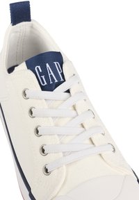 Witte canvas sneakers met een marineblauwe tong waarop het "GAP" logo staat, witte veters en een rubberen zool met een subtiele rode stripe accent.