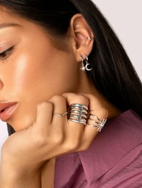 Gioielli d'argento con orecchini a cerchio decorati con charm a forma di luna e stella, anelli impilati e una fascia testurizzata, posati su una mano che si appoggia a una camicia viola.