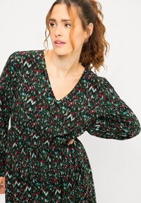 Vestido de manga larga con escote en V, que presenta un patrón multicolor en verde, rojo y azul, cintura ceñida y una falda fluida.
