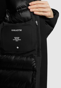 Juodų izoliacijos paltų vidus su siuvinėta tekstūra, pasižymi audinio etikete su užraša "KHUJOTM" ir "AUTENTIŠKAS BRANDS ĮSTEIGTAS 2001". Matoma gamybos kišenė su užtrauktuku.