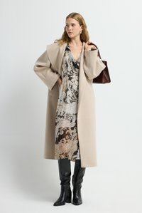 Donna che indossa un cappotto di lana beige sopra un vestito a fantasia, stivali neri fino al ginocchio, con una borsa marrone a tracolla, in piedi contro uno sfondo bianco.