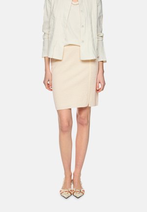 Frau trägt eine maßgeschneiderte cremefarbene Jacke, einen passenden knielangen Rock mit Frontschlitz und spitze High Heels mit Schnallendetail.