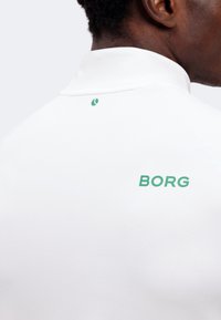 Άνδρας που φοράει λευκό πουκάμισο με γιακά και πράσινο κείμενο "BORG" στην άνω πλάτη και μικρό πράσινο λογότυπο κοντά στον γιακά.