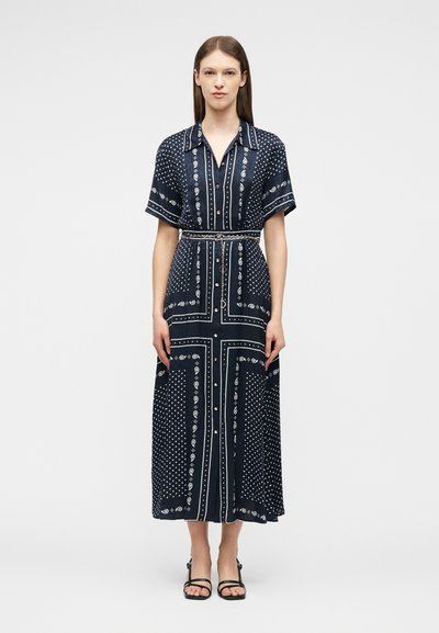 Γυναίκα που στέκεται φορώντας ένα μεσαίου μήκους φόρεμα σε ναυτικό μπλε χρώμα με λευκά σχέδια paisley και πουά, κοντά μανίκια, κουμπιά μπροστά και λεπτή ζώνη στη μέση.