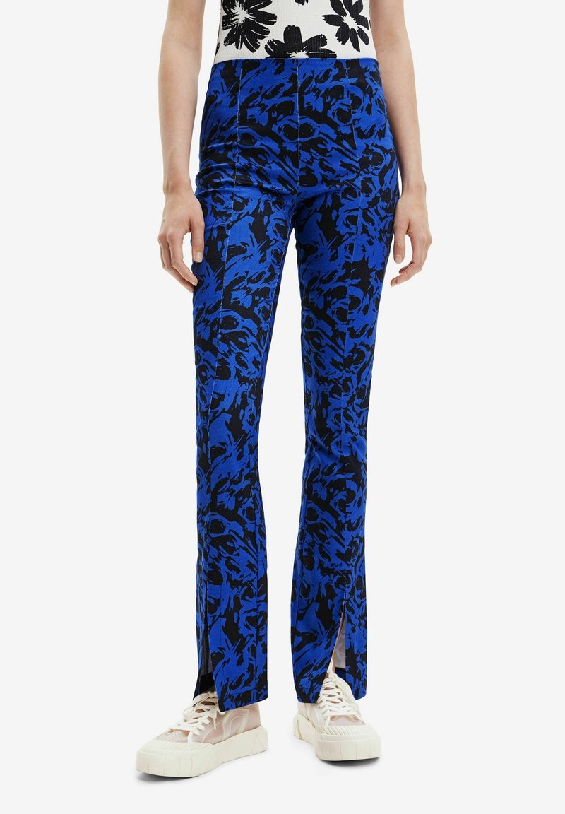 Pantalon slim ajusté à motif bleu et noir avec coutures frontales et fentes aux chevilles, porté avec des baskets plateformes blanches et un haut blanc imprimé floral noir.