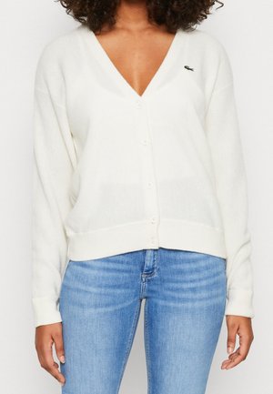 Kvinde iført en hvid knappet V-hals cardigan med et lille grønt logo på brystet og lyseblå jeans, hænderne afslappet langs siderne.
