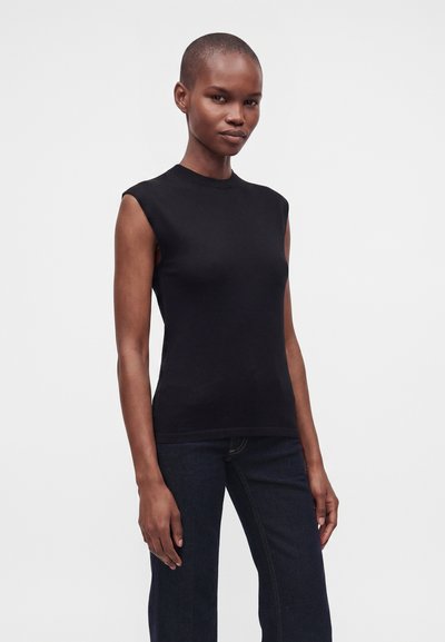 Γυναίκα με κοντά μαλλιά που φοράει μαύρο αμάνικο τοπ και σκούρο τζιν, στέκεται μπροστά σε απλό φωτεινό φόντο.