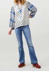 Blusa bianca con motivi floreali blu e maniche a palloncino, abbinata a jeans a zampa d'elefante azzurri e zoccoli di legno rossi con tacco.