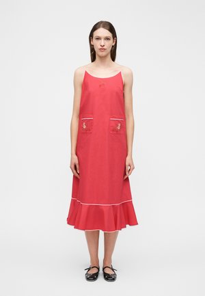 Γυναίκα που φοράει αμάνικο κόκκινο midi φόρεμα με κεντήματα λουλουδιών στις τσέπες και βολάν στο τελείωμα, συνδυασμένο με μαύρα παπούτσια Mary Jane.