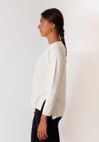 Maglione bianco a maglia con maniche lunghe, aperture laterali e scollatura a girocollo. Il tessuto ha una consistenza liscia e una leggera lavorazione a coste sulle spalle.