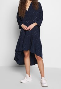 Marinblå klänning med V-ringning, långa ärmar och en lager-på-lager, asymmetrisk nederdel. Har texturerade prickar och mjukt tyg, kombinerad med vita sneakers.