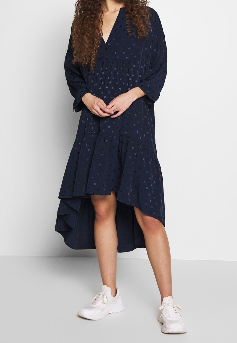 Marinblå klänning med V-ringning, långa ärmar och en lager-på-lager, asymmetrisk nederdel. Har texturerade prickar och mjukt tyg, kombinerad med vita sneakers.