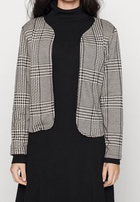 Svart och vit houndstooth-mönstrad cardigan med öppen framsida, långa ärmar och mjuk textur, bärs över en svart ribbad klänning.