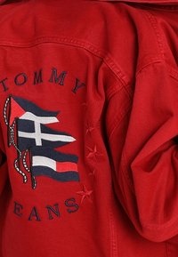 Veste en denim rouge avec logo et drapeaux brodés, et accents en étoile. Des coutures distinctives mettent en valeur la texture et les détails du tissu.