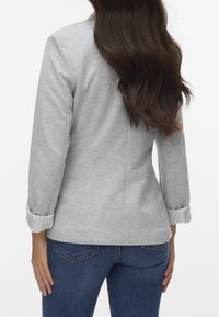 Žena s dlouhými hnědými vlasy, oblečená v světle šedém blejzru s vyhrnutými rukávy a tmavě modrých džínách, viděná zezadu.