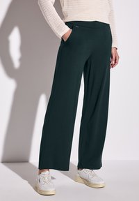 Pantaloni a gamba larga in tessuto verde scuro con una texture morbida, dotati di tasche laterali e una cintura comoda, abbinati a sneakers bianche.