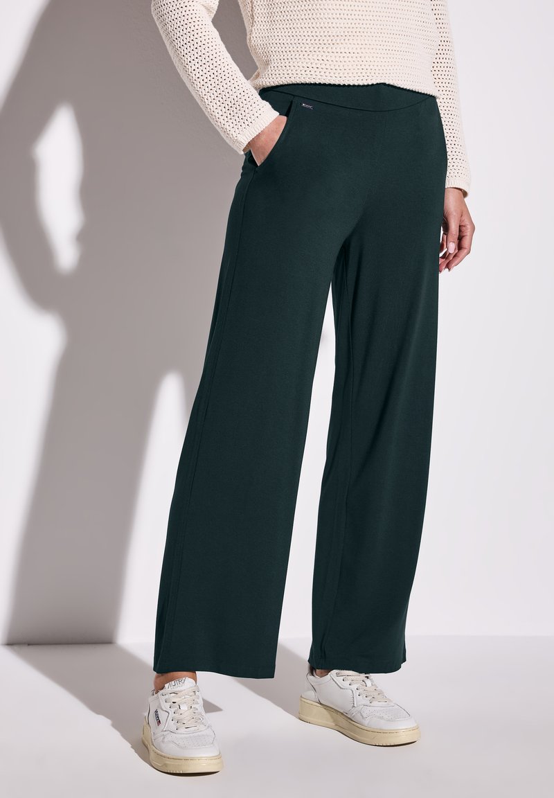 Pantaloni a gamba larga in tessuto verde scuro con una texture morbida, dotati di tasche laterali e una cintura comoda, abbinati a sneakers bianche.