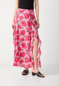 Jupe maxi florale en tissu rose avec des bords froncés, ornée de grandes fleurs rouges et de feuilles, associée à des sandales à talons blocs noires.