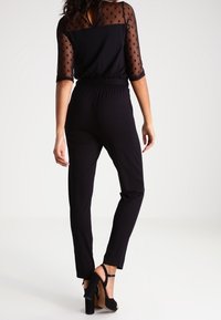 Zwarte jumpsuit met een doorzichtige top met polkadots en driekwartmouwen, een elastische tailleband en taps toelopende benen. Gepaard met zwarte sandalen met hakken.