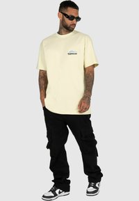 Geel katoenen T-shirt met korte mouwen, met een blauw en zwart graphic op de linkerborst. Gecombineerd met zwarte cargobroek en sneakers.