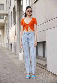 Top crop vermelho canelado com mangas curtas, jeans de perna reta de cintura alta azuis, chinelos de cor azul clara e uma mala de mão branca. Ambiente urbano.