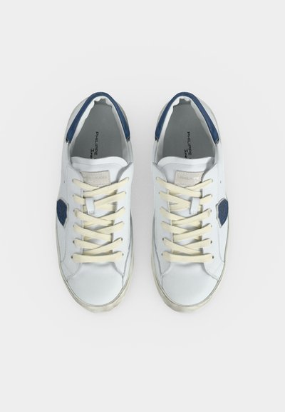 Paire de baskets blanches avec des lacets crème, des accents bleus au talon et des logos en forme de bouclier bleu sur les côtés, vues de dessus sur fond blanc.