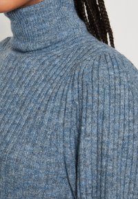 Pull bleu à col roulé côtelé, présentant une texture douce, un ajustement snug et des lignes verticales parcourant le tissu.