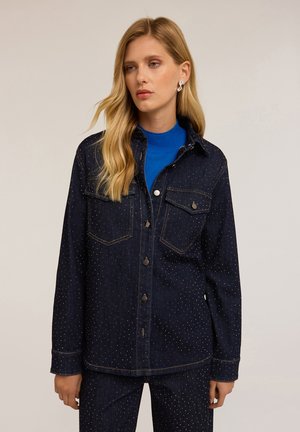 Mujer que lleva una chaqueta de mezclilla oscura y pantalones con pequeños puntos dorados, combinados con un suéter de cuello alto azul brillante y pendientes de plata.