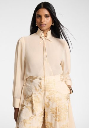 Frau mit langen schwarzen Haaren trägt eine beige Bluse mit hohem Kragen und Schleife sowie cremefarbene, weit geschnittene Hosen mit goldenem Blumenmuster, die Hände in den Taschen.