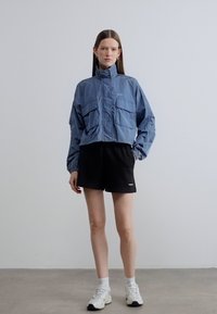 Μπλε μπουφάν με κοντό μήκος, δύο μπροστινές τσέπες, ελαστικά μανίκια και ψηλό γιακά, σε συνδυασμό με μαύρο σορτς και λευκά αθλητικά παπούτσια. Απλός σχεδιασμός.