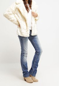 Faux fur krämfärgad jacka med bred krage, ihopparad med denimjeans och bruna ankelstövlar. Tyget har en texturerad och lyxig finish.