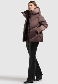 Kvinne iført en brun polstret jakke med høy krage, svarte bukser og svarte kileboots, som står med den ene hånden i lommen.