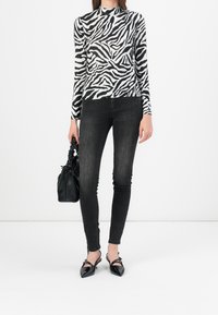 Mujer vestida con una blusa de manga larga de estampado de cebra blanco y negro, jeans ajustados negros, zapatos de charol negros de punta, sosteniendo un bolso negro arrugado.