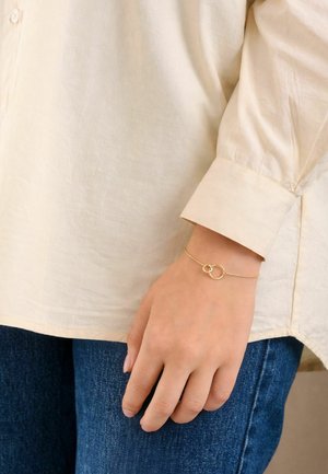 Roka z nežno zlato zapestnico s dvema medsebojno povezanima prstanoma, ki počiva ob strani, oblečena v kremasto dolgoročno majico in modre kavbojke.