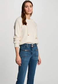 Sudadera de felpa crema con cordones, combinada con jeans azules que incluyen elementos decorativos y bolsillos frontales. Textura suave y corte relajado.