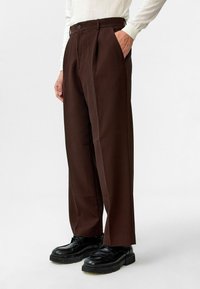 Bruine geplooide broek met verticale ribbels, voorzien van twee zijzakken en een knoopsluiting, gedragen met zwarte veterschoenen.