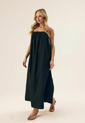 Schwarzes, ärmelloses Maxikleid mit gerafftem Ausschnitt, fließendem Stoff und knöchellanger Passform. Kombiniert mit tanfarbenen, minimalistischen Sandalen.