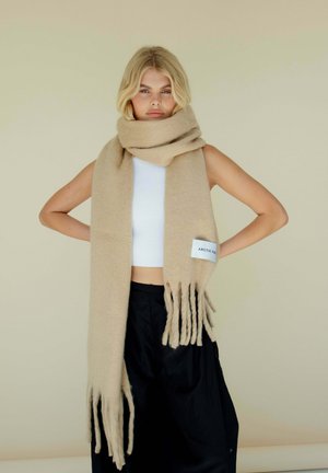 Écharpe en laine beige avec des extrémités à franges, de forme rectangulaire et avec une étiquette en tissu. Portée autour du cou sur un fond neutre.