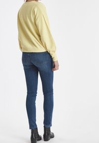 Gul stickad tröja med lösa ärmar, kombinerad med smala mörkblå jeans. Modellen har på sig svarta ankelboots.