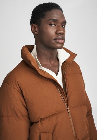 Doudoune marron avec un col montant, fermeture éclair à l'avant et poches latérales. Le matériau semble lisse et légèrement texturé avec une silhouette moderne.