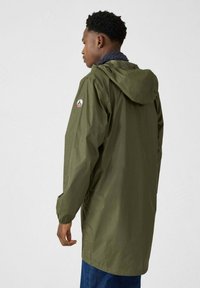 Veste imperméable vert olive avec capuche, manches longues et poches latérales. Présente un patch logo sur la manche gauche. Texture lisse, coupe légèrement ample.