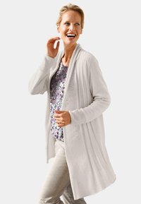 Lichtbeige lange cardigan van gebreide stof, met ribbeltextuur, open voorzijde en zijplooien. Draagt over een top met bloemenpatroon.