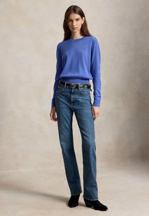 Ung kvinde står iført en blå bluse med lange ærmer, højttaljede blå jeans med sort bælte og sorte sko med hæl.
