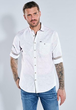 Hellblaues Button-down-Hemd mit einem subtilen Muster, hochgekrempelte Ärmel und einer Brusttasche, kombiniert mit blauen Jeans. Verfügt über braune Knöpfe.