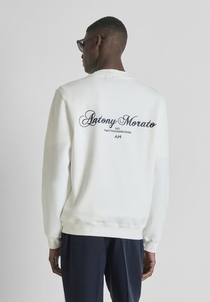 Man draagt een wit sweatshirt met de tekst "Antony Morato" op de achterkant, gecombineerd met een donkere broek, staat met de rug naar de camera op een effen achtergrond.