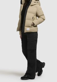 Beżowa kurtka puchowa z kapturem i czarnymi akcentami, zestawiona z czarnymi spodniami cargo z wieloma kieszeniami i czarnymi sneakersami.