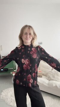 Junge Frau mit blondem Haar, die eine schwarze Bluse mit Blumenmuster und schwarze Hose trägt, steht mit ausgebreiteten Armen in einem hellen Wohnzimmer.
