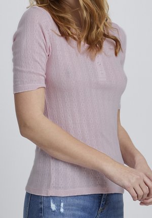 Femme aux cheveux ondulés portant un haut en maille à manches courtes de couleur rose clair avec un motif, orné de trois petits boutons, et un jean bleu déchiré.