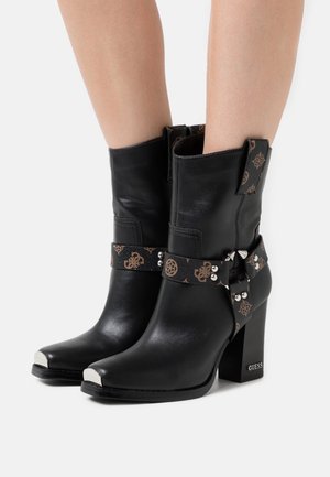 Sorte læderstøvler til midt på læggen med blokhæle, mærkede metal-tåsko, dekorative mønstrede remme med nitter, båret af en person.